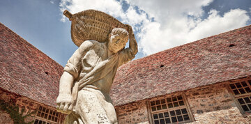 Statue aus Stein mit einem Mönch in einem Kloster im Burgund, der eine Kiepe mit vermutlich Traiben trägt.